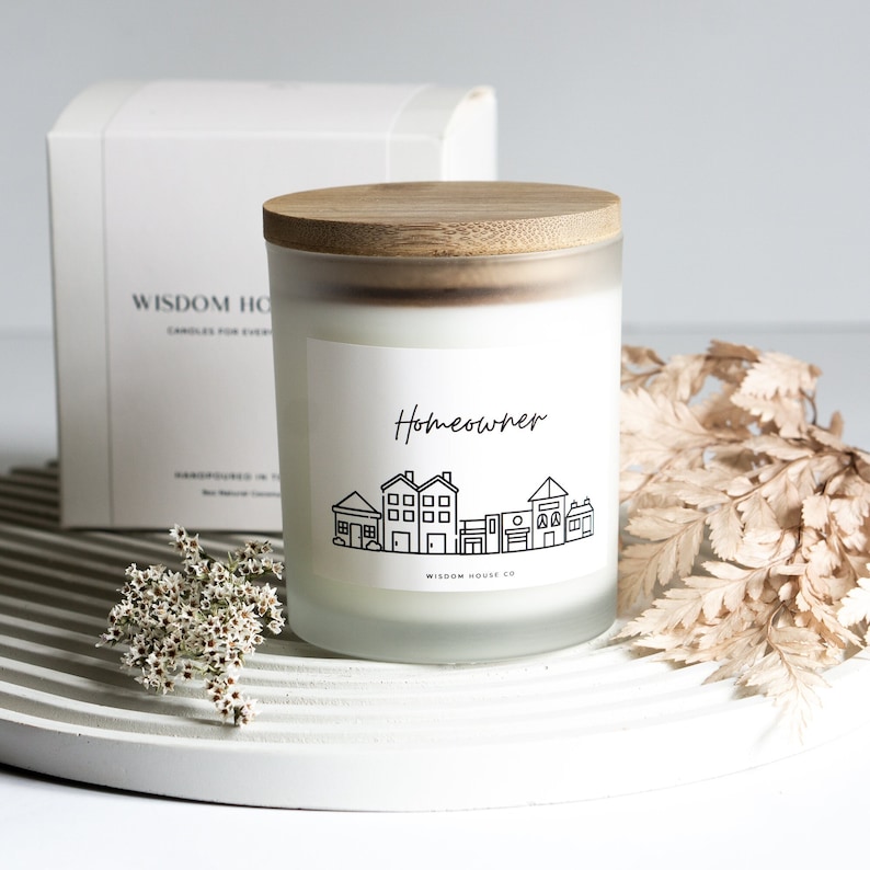 Op de afbeelding: Een witte, gematteerde glazen kaars met een houten deksel. De kaars heeft een zwart-wit label met de tekst "Homeowner" en een afbeelding van een rij huizen. De kaars staat op een wit rond oppervlak.