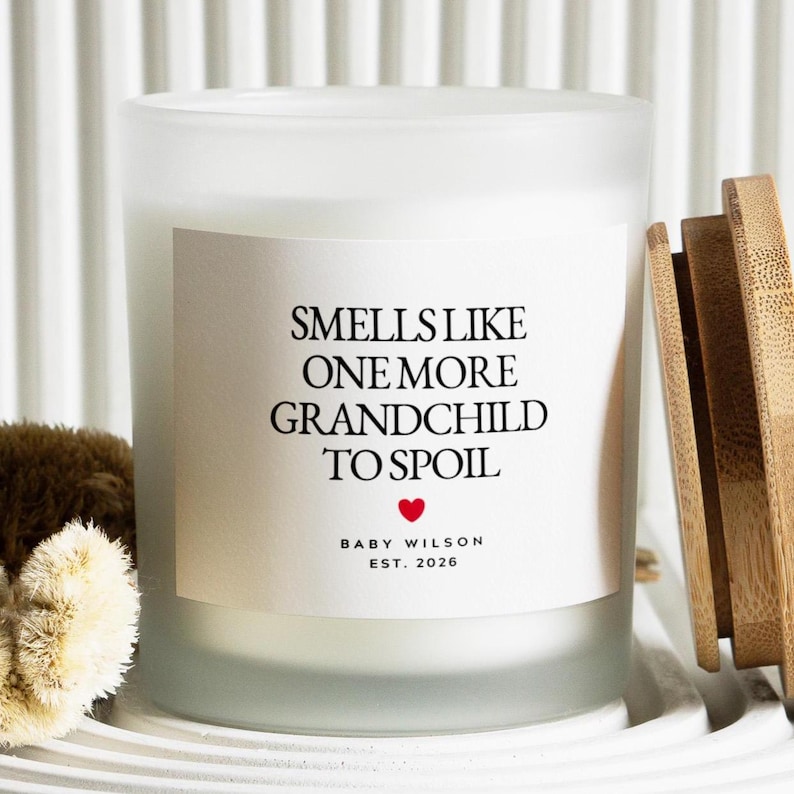 Pu&ograve; includere: Una candela bianca in un barattolo di vetro smerigliato con coperchio in legno. L'etichetta recita "Smells Like One More Grandchild To Spoil" con un cuore rosso e "Baby Wilson Est. 2026".