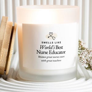 May include: A lit, frosted glass candle with a wooden lid, on a white tray. The candle's label says "Smells Like World's Best Nurse Educator" with a stethoscope heart logo. A sprig of white flowers is next to the candle. The candle is a home decor item.