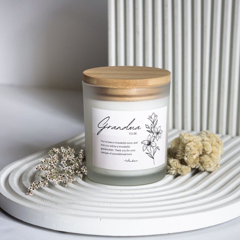 Puede incluir: Un tarro de vela de vidrio blanco esmerilado con una tapa de madera. El tarro tiene una etiqueta en blanco y negro con el texto "Grandma to be" y un dise&ntilde;o floral. El texto dice "You've been a wonderful mom, and now you will be a wonderful grandmother. Thank you for your example of unconditional love. - Amber"