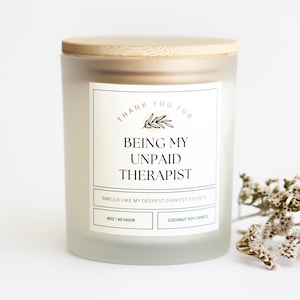 May include: A white frosted glass candle jar with a bamboo lid. The label reads "Thank you for being my unpaid therapist. Smells like my deepest darkest secrets." The candle is 8 oz and has a 45-hour burn time. It is a coconut soy candle.