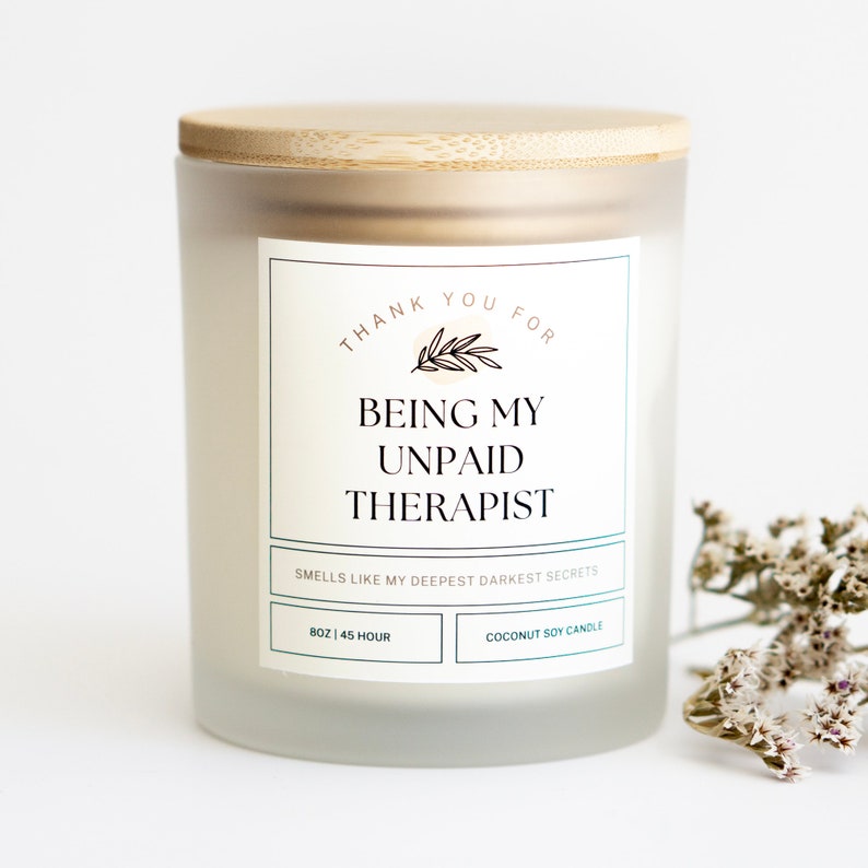 May include: A white frosted glass candle jar with a bamboo lid. The label reads "Thank you for being my unpaid therapist. Smells like my deepest darkest secrets." The candle is 8 oz and has a 45-hour burn time. It is a coconut soy candle.