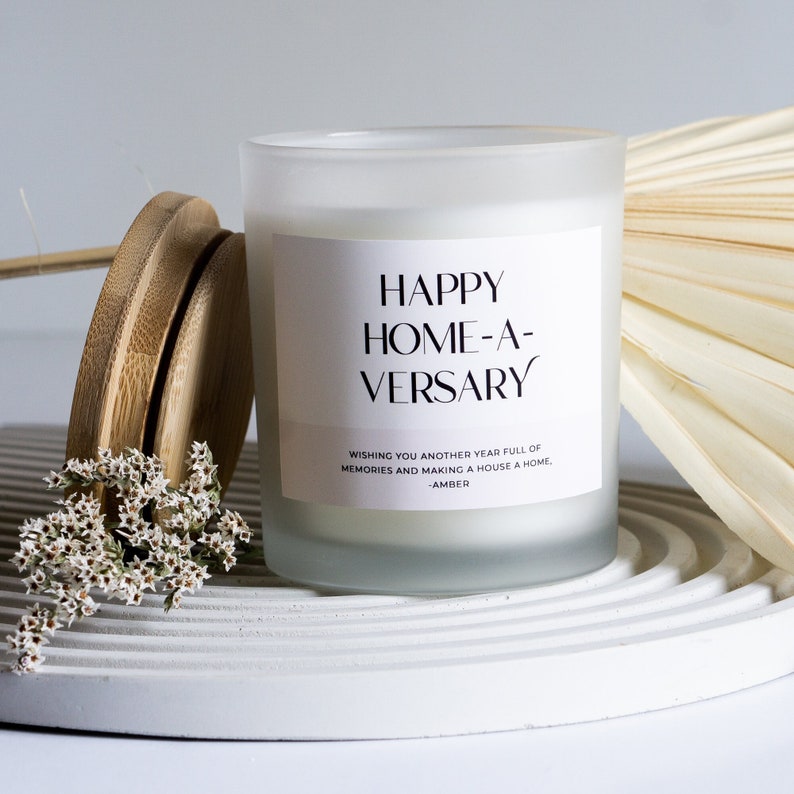 May include: White frosted glass candle jar with a white label that reads "HAPPY HOME-A-VERSARY WISHING YOU ANOTHER YEAR FULL OF MEMORIES AND MAKING A HOUSE A HOME, -AMBER".