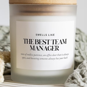 Puede incluir: Una vela en frasco de vidrio esmerilado con tapa de madera clara y etiqueta blanca. La etiqueta dice: SMELLS LIKE THE BEST TEAM MANAGER, notes of endless patience, an office door that is always open, and knowing someone always has your back.