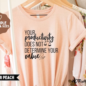 May include: A peach colored t-shirt with black text that reads "Your productivity does not determine your value" with a heart and a scale graphic.