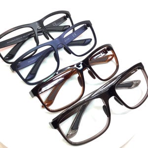 May include: Four pairs of eyeglasses in various colors, including black, navy blue, and brown. The glasses have rectangular frames and are arranged in a diagonal line, showcasing their different styles and designs.