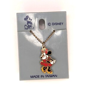 May include: A gold-colored necklace with a Minnie Mouse pendant. The pendant features Minnie in a red dress, black shoes, and a red bow. The necklace is attached to a gray cardboard backing with the Disney logo and the text "MADE IN TAIWAN".