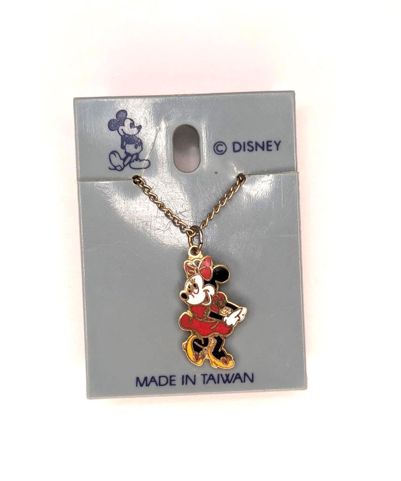 May include: A gold-colored necklace with a Minnie Mouse pendant. The pendant features Minnie in a red dress, black shoes, and a red bow. The necklace is attached to a gray cardboard backing with the Disney logo and the text "MADE IN TAIWAN".