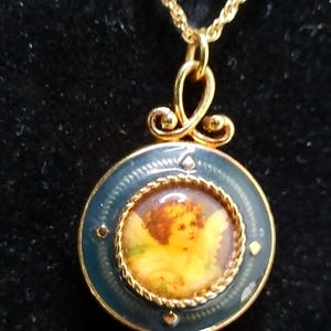 May include: Gold-tone necklace with a pendant featuring a cherub painting. The pendant has a blue enamel border and a decorative bail. The necklace has a delicate chain and a cluster of clear crystals at the bottom of the pendant.