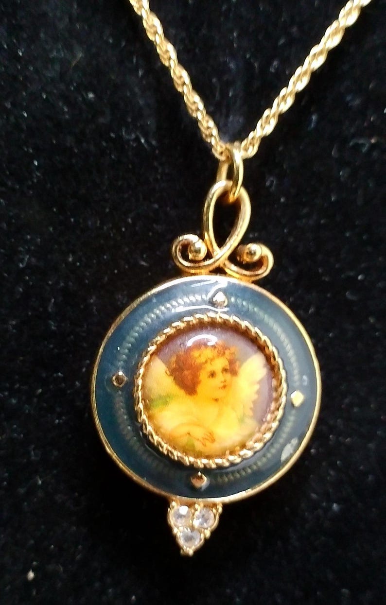 May include: Gold-tone necklace with a pendant featuring a cherub painting. The pendant has a blue enamel border and a decorative bail. The necklace has a delicate chain and a cluster of clear crystals at the bottom of the pendant.