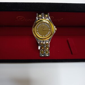 May include: A gold and silver-toned Donna Ricco wristwatch with a gold face and diamond accents. The watch is displayed in a red velvet box with the brand name printed on the inside. The watch band is a combination of gold and silver links.