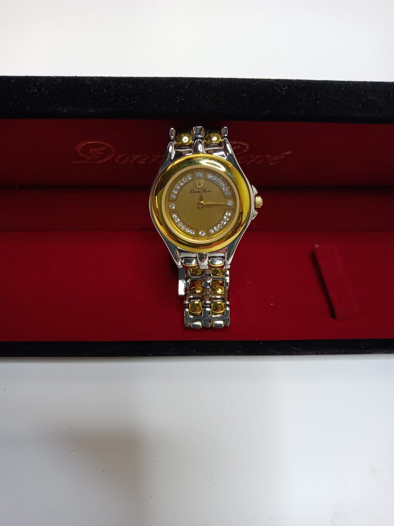 May include: A gold and silver-toned Donna Ricco wristwatch with a gold face and diamond accents. The watch is displayed in a red velvet box with the brand name printed on the inside. The watch band is a combination of gold and silver links.