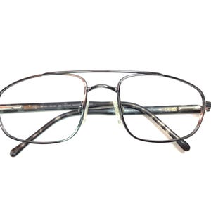 May include: Aviator-style eyeglasses with a thin, dark metal frame and clear lenses. The temples feature a tortoiseshell pattern. The glasses have a double bridge and a classic, timeless design.