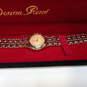 May include: A Donna Ren&eacute; wristwatch in a red velvet box. The watch has a gold-tone bezel and band with a light-colored face. The watch face has gold-tone hands and hour markers. The brand name is in gold script on the box.