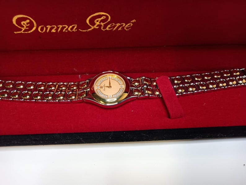 May include: A Donna Ren&eacute; wristwatch in a red velvet box. The watch has a gold-tone bezel and band with a light-colored face. The watch face has gold-tone hands and hour markers. The brand name is in gold script on the box.