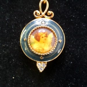 May include: Gold-toned pendant with a circular, teal enamel frame. The center features a painted cherub image. The pendant has a decorative bail and a teardrop-shaped cluster of clear crystals at the bottom. A delicate, vintage-style jewelry piece.