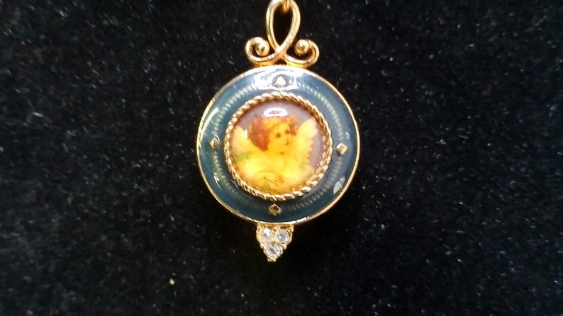 May include: Gold-toned pendant with a circular, teal enamel frame. The center features a painted cherub image. The pendant has a decorative bail and a teardrop-shaped cluster of clear crystals at the bottom. A delicate, vintage-style jewelry piece.