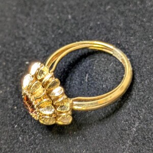 May include: A gold-colored ring with a cluster of textured, petal-like shapes on one side. The ring has a double band design and a small, dark-colored stone is visible within the cluster. The ring is set against a dark background.