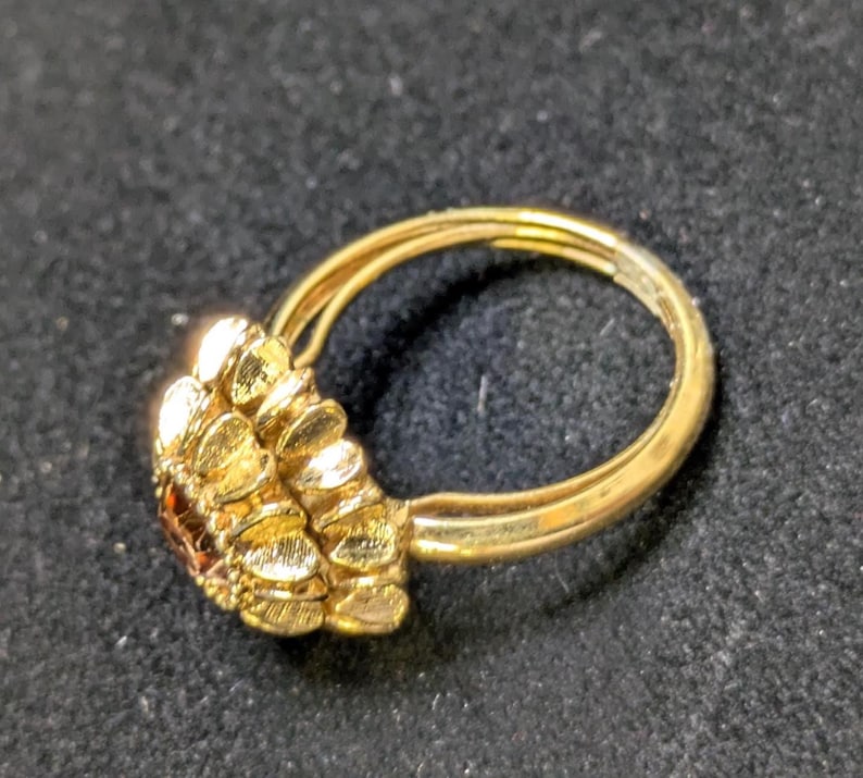 May include: A gold-colored ring with a cluster of textured, petal-like shapes on one side. The ring has a double band design and a small, dark-colored stone is visible within the cluster. The ring is set against a dark background.