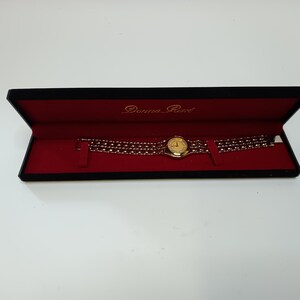 May include: A gold-toned Donna Reed wristwatch with a round face and a multi-strand band, presented in a black box with a red interior. The watch face is gold, and the band is made of small, connected links.