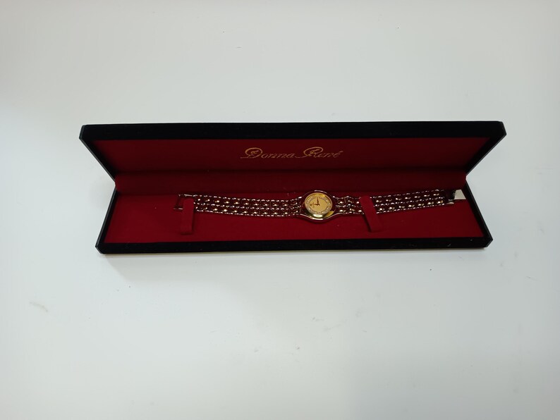 May include: A gold-toned Donna Reed wristwatch with a round face and a multi-strand band, presented in a black box with a red interior. The watch face is gold, and the band is made of small, connected links.