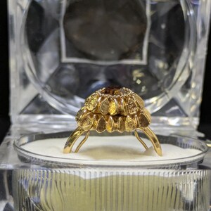 May include: A gold ring with a floral design, set with a central brown gemstone. The ring is displayed in a clear, geometric jewelry box with a white interior, against a black background.