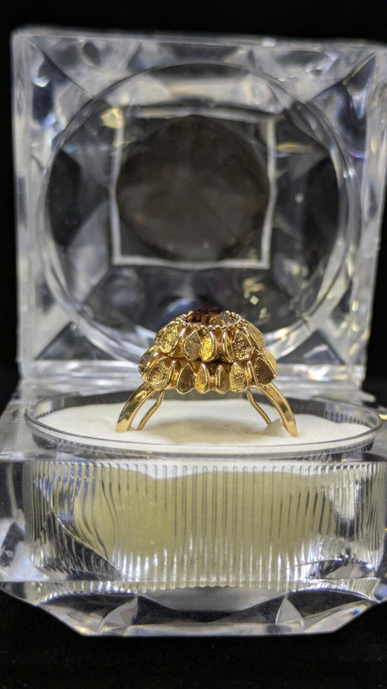 May include: A gold ring with a floral design, set with a central brown gemstone. The ring is displayed in a clear, geometric jewelry box with a white interior, against a black background.