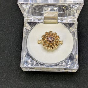 May include: A gold-colored ring with a central, round, light brown gemstone, surrounded by a floral design. The ring is displayed in a clear, hinged plastic box with a white interior. The box is open, revealing the ring.
