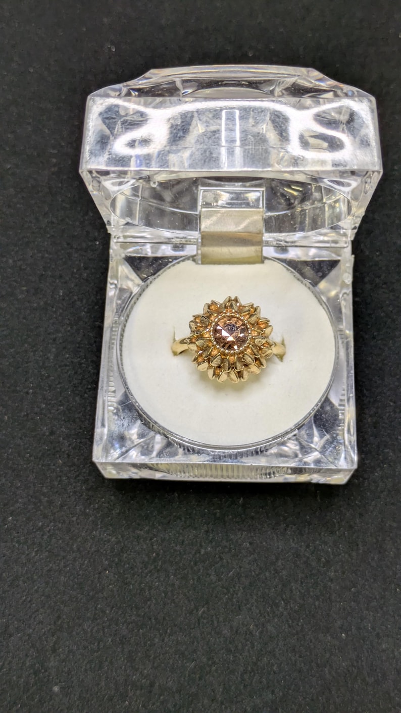 May include: A gold-colored ring with a central, round, light brown gemstone, surrounded by a floral design. The ring is displayed in a clear, hinged plastic box with a white interior. The box is open, revealing the ring.