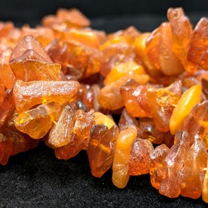 May include: A necklace composed of amber stones, displayed in a close-up shot. The amber pieces are of varying sizes and shapes, exhibiting a warm, translucent orange hue. The necklace is strung together, presenting a natural, organic aesthetic.