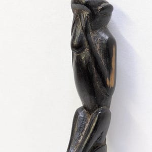 Pode incluir: Uma estatueta de madeira esculpida de uma figura sentada com as mãos cobrindo o rosto. A escultura marrom escuro tem um acabamento liso e polido e é montada em uma base retangular. A forma da figura é estilizada e alongada.
