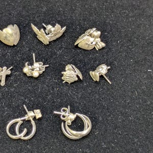 Puede incluir: Una colección de pendientes plateados con diversos diseños florales y abstractos. Los pendientes incluyen estilos de botón y aro, algunos con detalles de perlas. Las piezas se exhiben sobre una superficie oscura y texturizada.