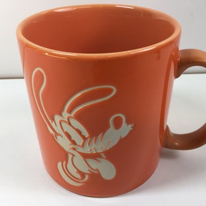 Peut inclure: Une tasse en céramique orange avec une silhouette blanche d'une souris de dessin animé. La souris est dans une pose de saut avec ses bras et ses jambes tendus.