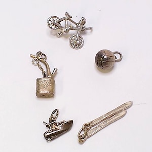 May include: A selection of silver charms, featuring a bicycle, a volleyball, a boat, a bong, and a pair of scissors. The charms are detailed and possibly made of metal, with small loops for attaching to a bracelet or necklace.