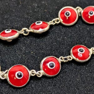 May include: Silver bracelet with red enamel eye charms, black pupils, and white highlights. The bracelet is linked with silver and has a lobster clasp. The eye charms are approximately 2 cm in diameter.