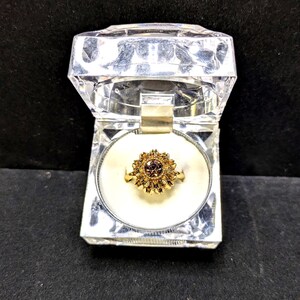 May include: A gold-colored ring with a flower-like design, featuring a central dark stone surrounded by smaller petals. The ring is displayed in a clear, faceted jewelry box with a white interior.
