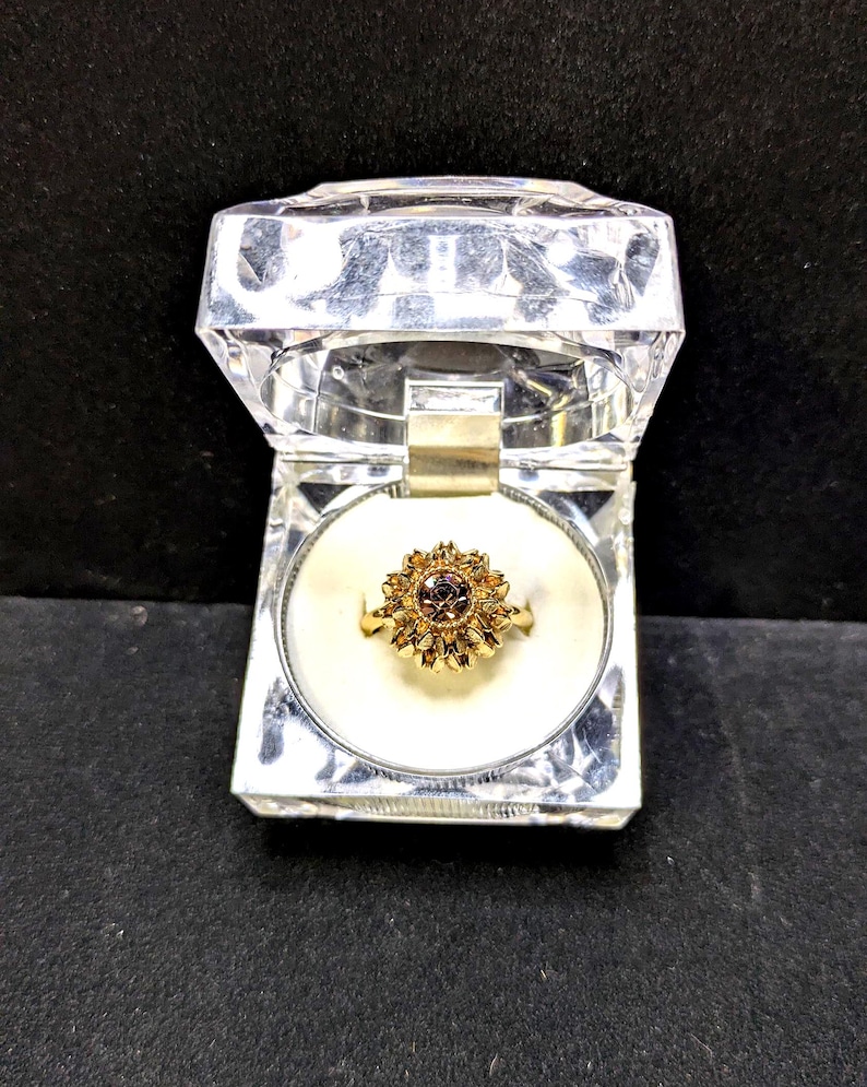 May include: A gold-colored ring with a flower-like design, featuring a central dark stone surrounded by smaller petals. The ring is displayed in a clear, faceted jewelry box with a white interior.