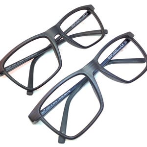 May include: Two pairs of matte black eyeglasses with rectangular frames. The glasses have a modern design with thick rims and temples. The lenses are clear, and the frames appear to be made of a durable material. The glasses are displayed on a white background.