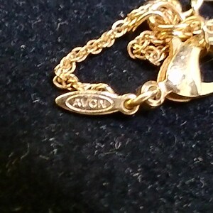 May include: Close-up of a gold-colored necklace clasp and chain. The clasp is a lobster claw style, and the chain is delicate. The pendant tag reads "AVON". The jewelry is set against a dark background.