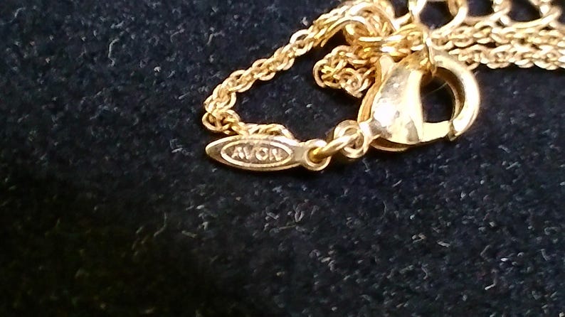 May include: Close-up of a gold-colored necklace clasp and chain. The clasp is a lobster claw style, and the chain is delicate. The pendant tag reads "AVON". The jewelry is set against a dark background.