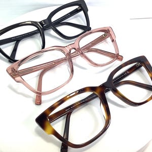 May include: Three pairs of eyeglasses with different colored frames: black, pink, and tortoise shell. All three pairs have a square shape with rounded corners.