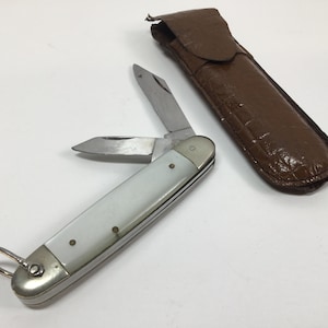 Puede incluir: Un cuchillo de bolsillo con mango blanco, dos hojas y un pequeño anillo en el extremo del mango. El cuchillo está abierto y las hojas están extendidas. El cuchillo está en una funda de cuero marrón.
