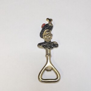 May include: A vintage bottle opener featuring a decorative bust of a man in a black hat with a red detail. The opener is made of gold-coloured metal and has a classic bottle-opening design.
