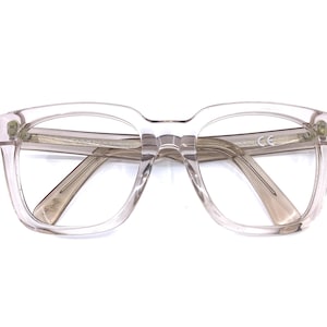 May include: Clear, square-framed eyeglasses with a light tint. The frames are transparent, showcasing the lenses and the subtle design of the temples. The glasses have a classic, slightly oversized shape.