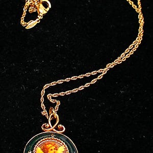 May include: Gold-toned necklace featuring a pendant with a painted cherub image. The pendant is round, framed in dark green enamel with a decorative border, and accented with small crystals. The chain is a delicate rope style.