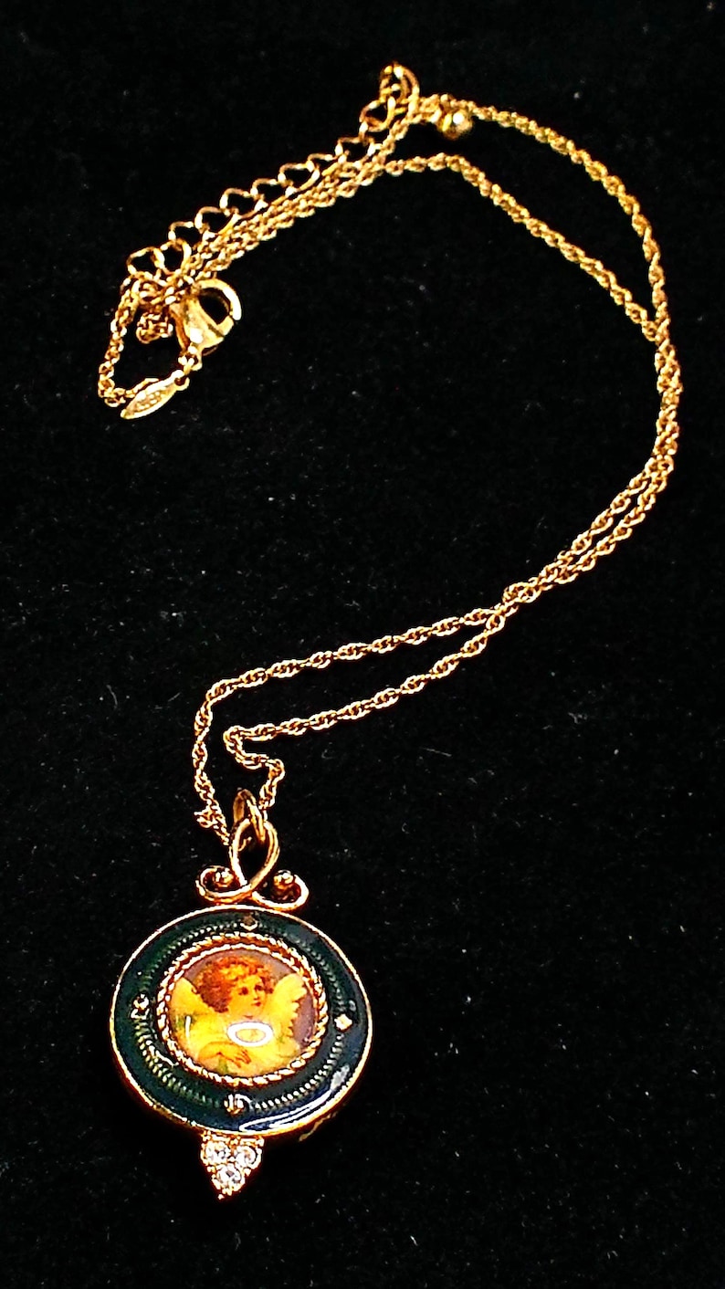 May include: Gold-toned necklace featuring a pendant with a painted cherub image. The pendant is round, framed in dark green enamel with a decorative border, and accented with small crystals. The chain is a delicate rope style.