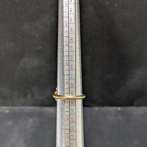 May include: A gold ring is placed on a ring sizer. The ring has a small, decorative element on top. The ring sizer is silver with black and red markings, indicating ring sizes. The background is a dark gray.