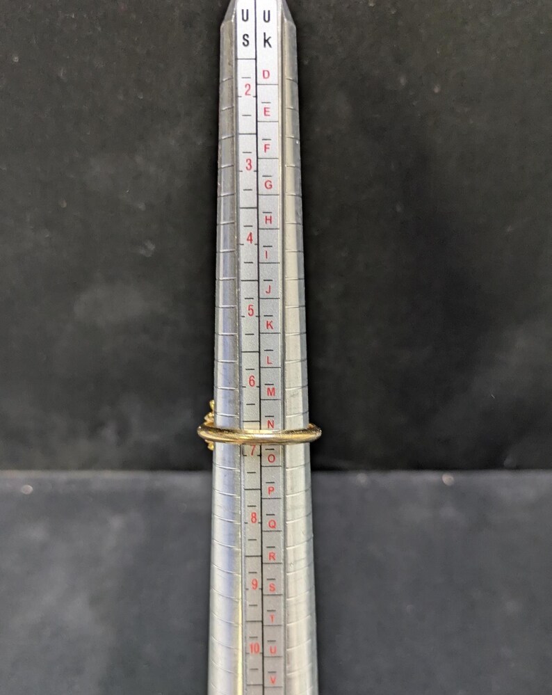 May include: A gold ring is placed on a ring sizer. The ring has a small, decorative element on top. The ring sizer is silver with black and red markings, indicating ring sizes. The background is a dark gray.