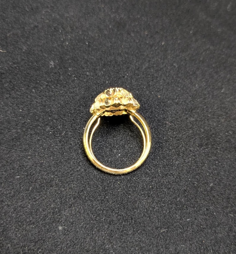 May include: A gold ring with a floral design. The ring features a double band and a cluster of small, textured petals forming the centerpiece. The ring is set against a dark background, highlighting its bright, shiny appearance.