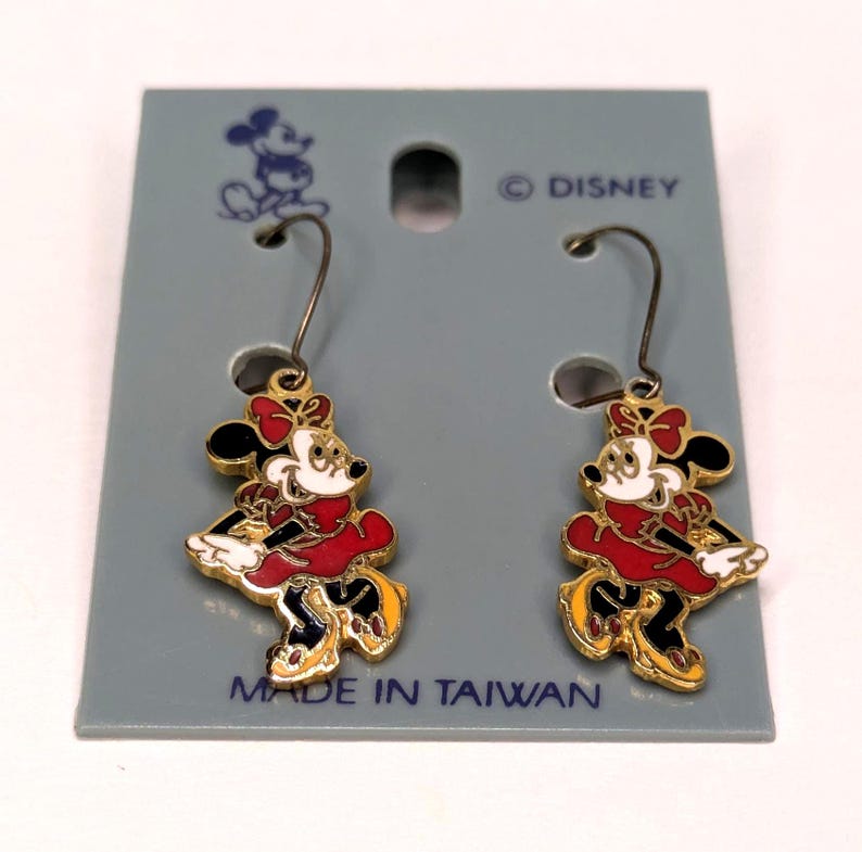 May include: A pair of Minnie Mouse dangle earrings. The earrings feature a gold-colored outline with black, red, white, and yellow enamel details. The earrings are attached to a gray card that reads "&copy; DISNEY" and "MADE IN TAIWAN."